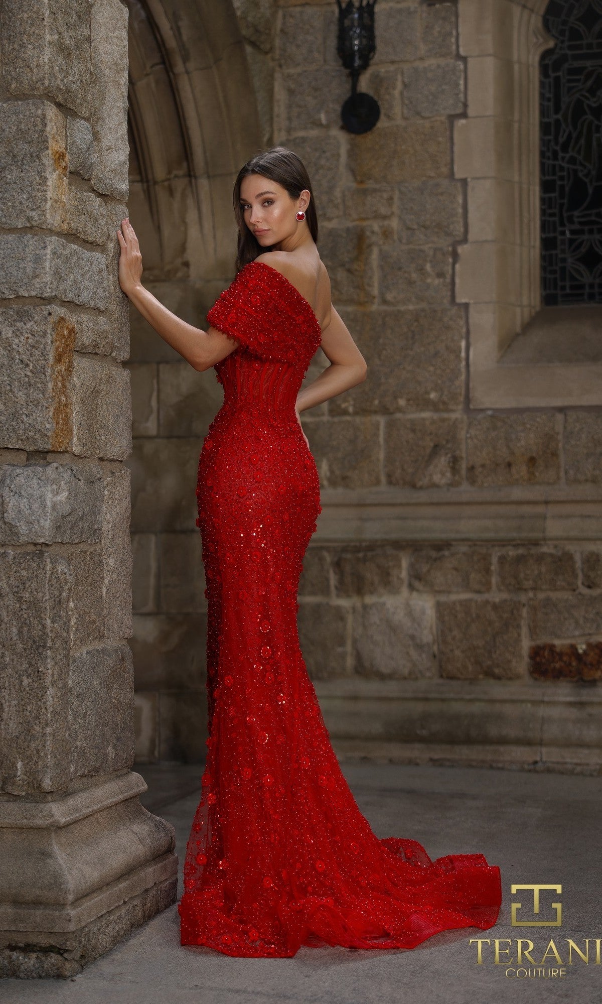 A woman stands by a stone wall in an elegant setting wearing the Terani 261GL6756 long formal dress, an off-the-shoulder, floor-length red gown with a fitted silhouette and intricate beaded tulle detailing.